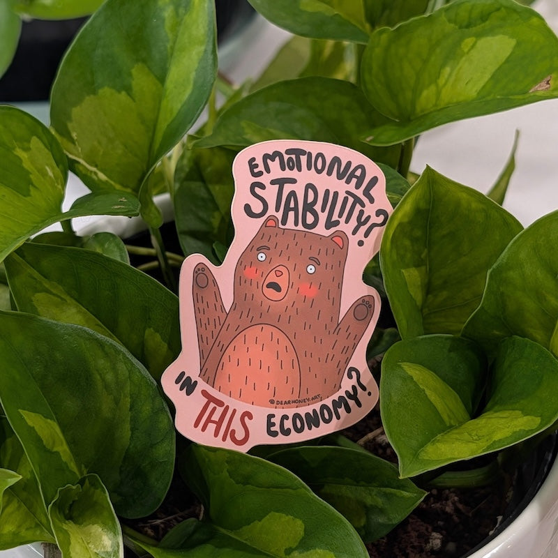 A die-cut sticker featuring a cartoon brown bear with the text 'EMOTIONAL STABILITY? IN THIS ECONOMY?' placed among green houseplant leaves.