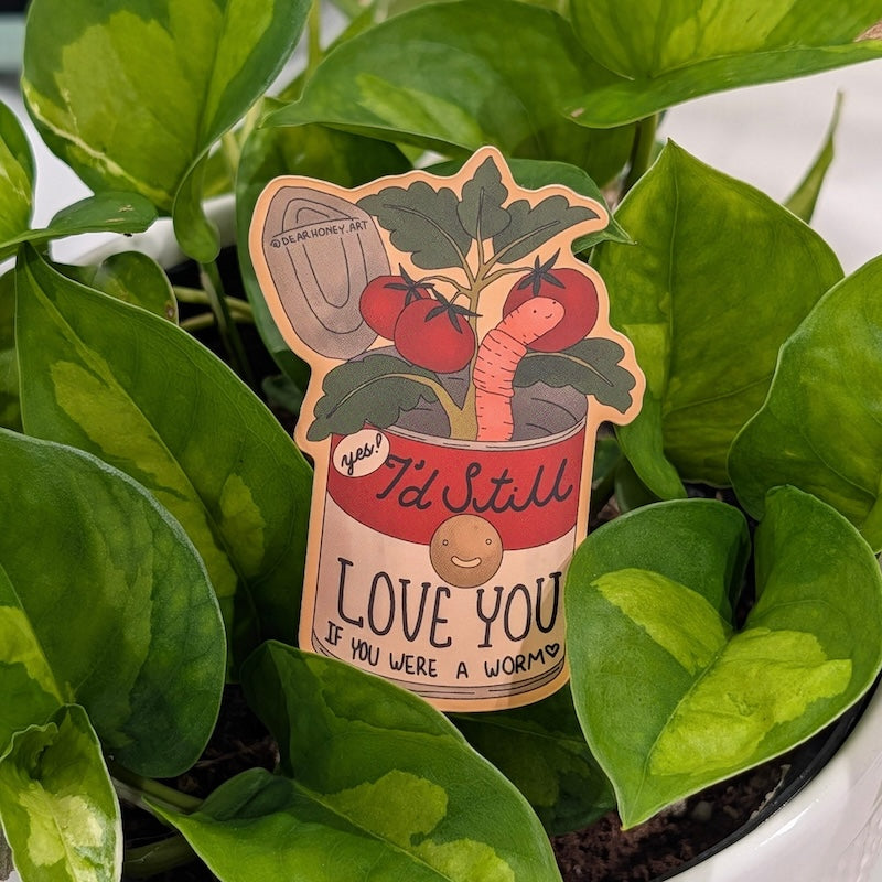 A die-cut sticker featuring a whimsical tomato illustration with the text 'I'd Still LOVE YOU IF YOU WERE A WORM', placed among green plant leaves.